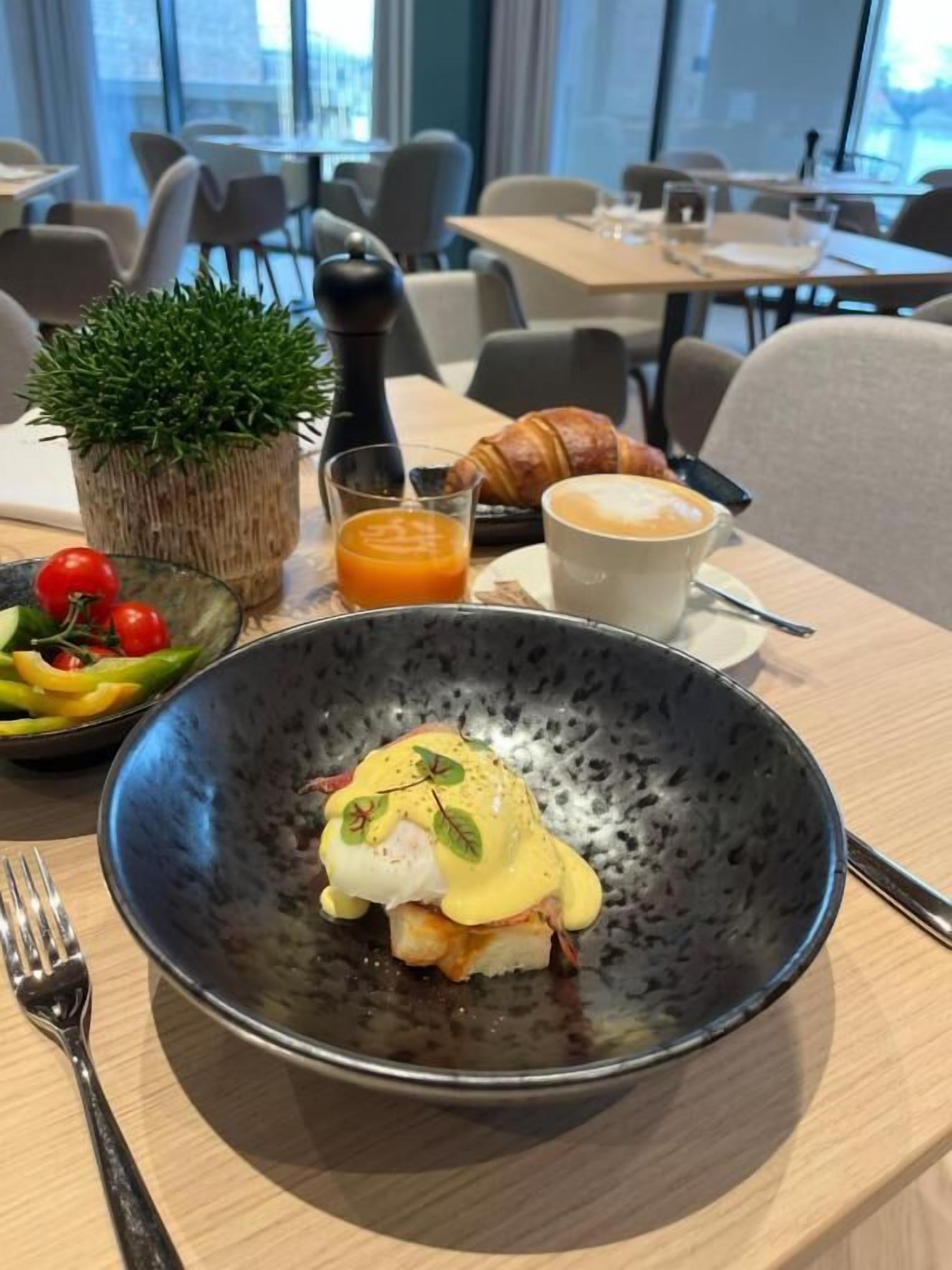 Una mesa de comedor con un desayuno que incluye huevos benedictinos, un croissant y un vaso de jugo de naranja.