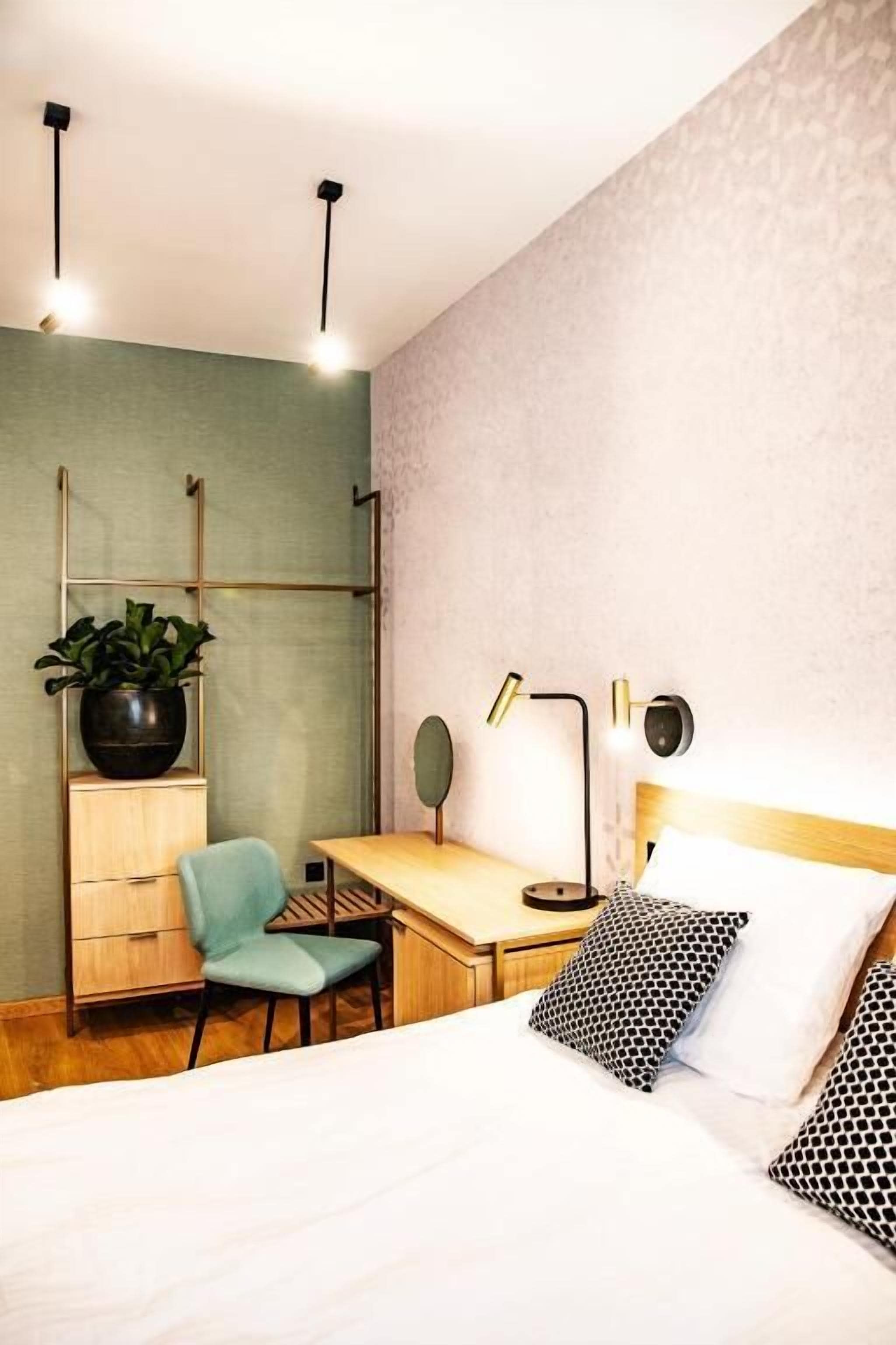 A bedroom with a bed, a desk, a chair, a potted plant, and a wall-mounted lamp.