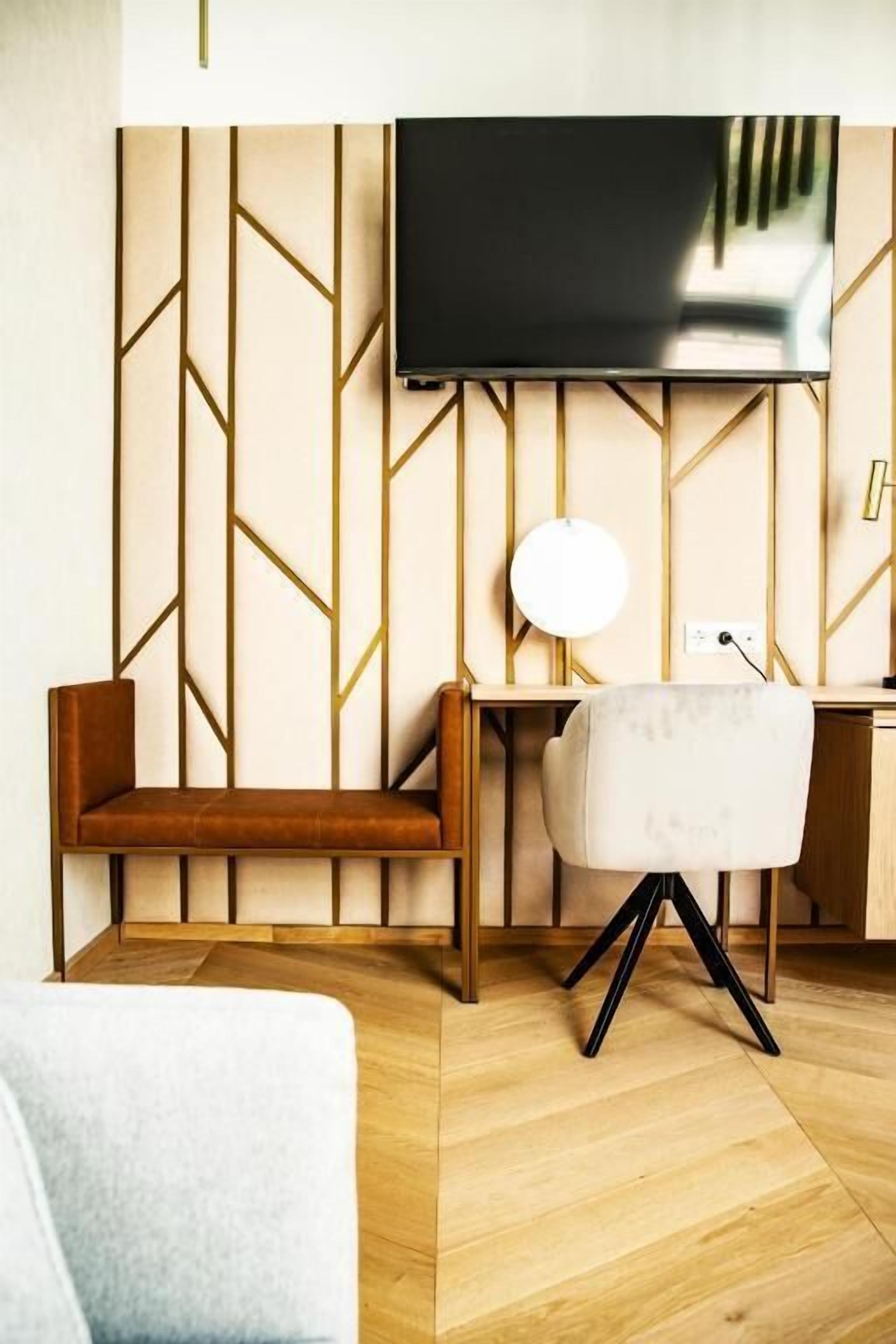 A modern room with a wooden floor, a sofa, a desk with a chair, a television mounted on the wall, and a lamp.