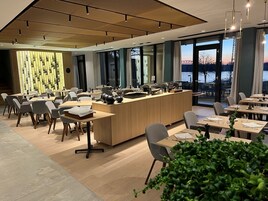 Uma zona de refeições moderna com buffet central, várias mesas e cadeiras, grandes janelas com vista para um corpo de água.