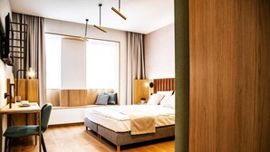 Una habitación de hotel moderna con piso de madera, un escritorio con silla, cama con almohadas, ventana con cortinas y un televisor instalado en la pared.
