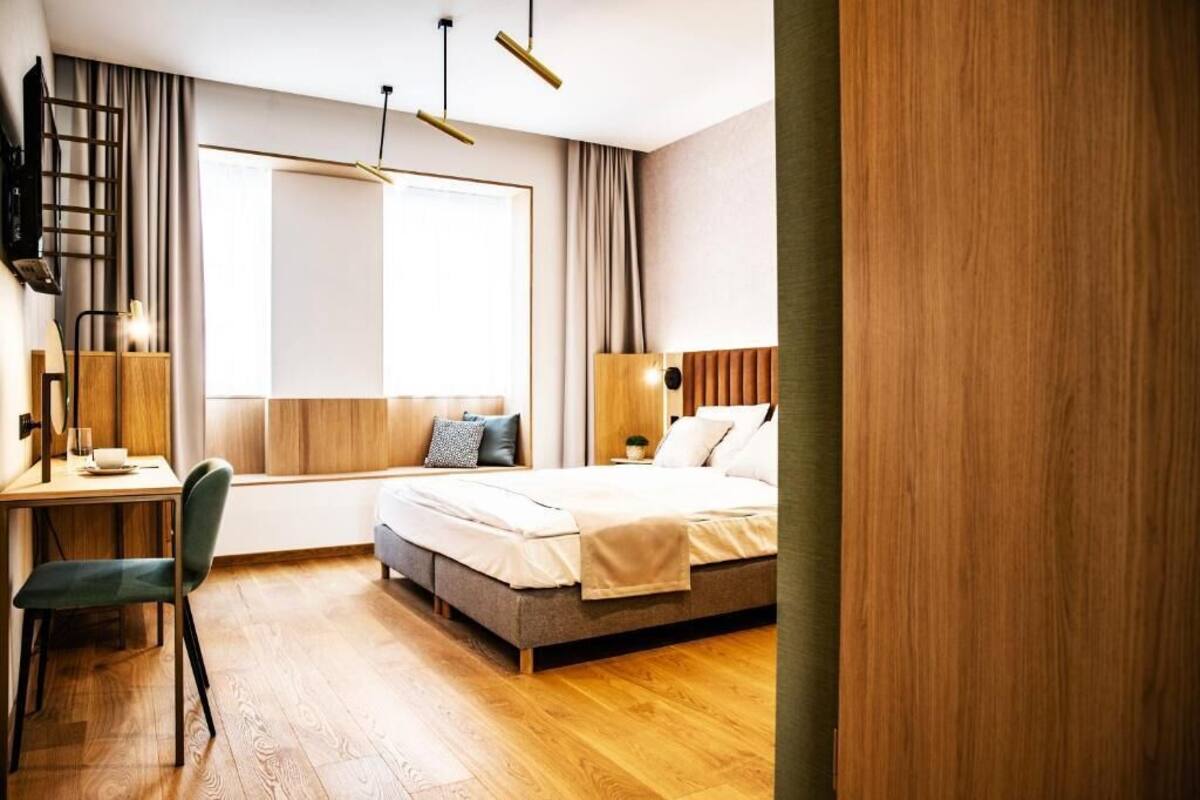 Una habitación de hotel moderna con piso de madera, un escritorio con silla, cama con almohadas, ventana con cortinas y un televisor instalado en la pared.