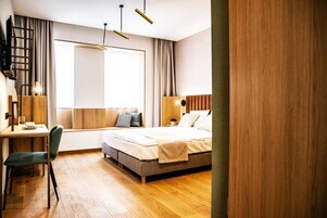 Quarto de hotel moderno com piso de madeira, uma escrivaninha com cadeira, uma cama com almofadas, uma janela com cortinas e uma televisão fixada na parede.
