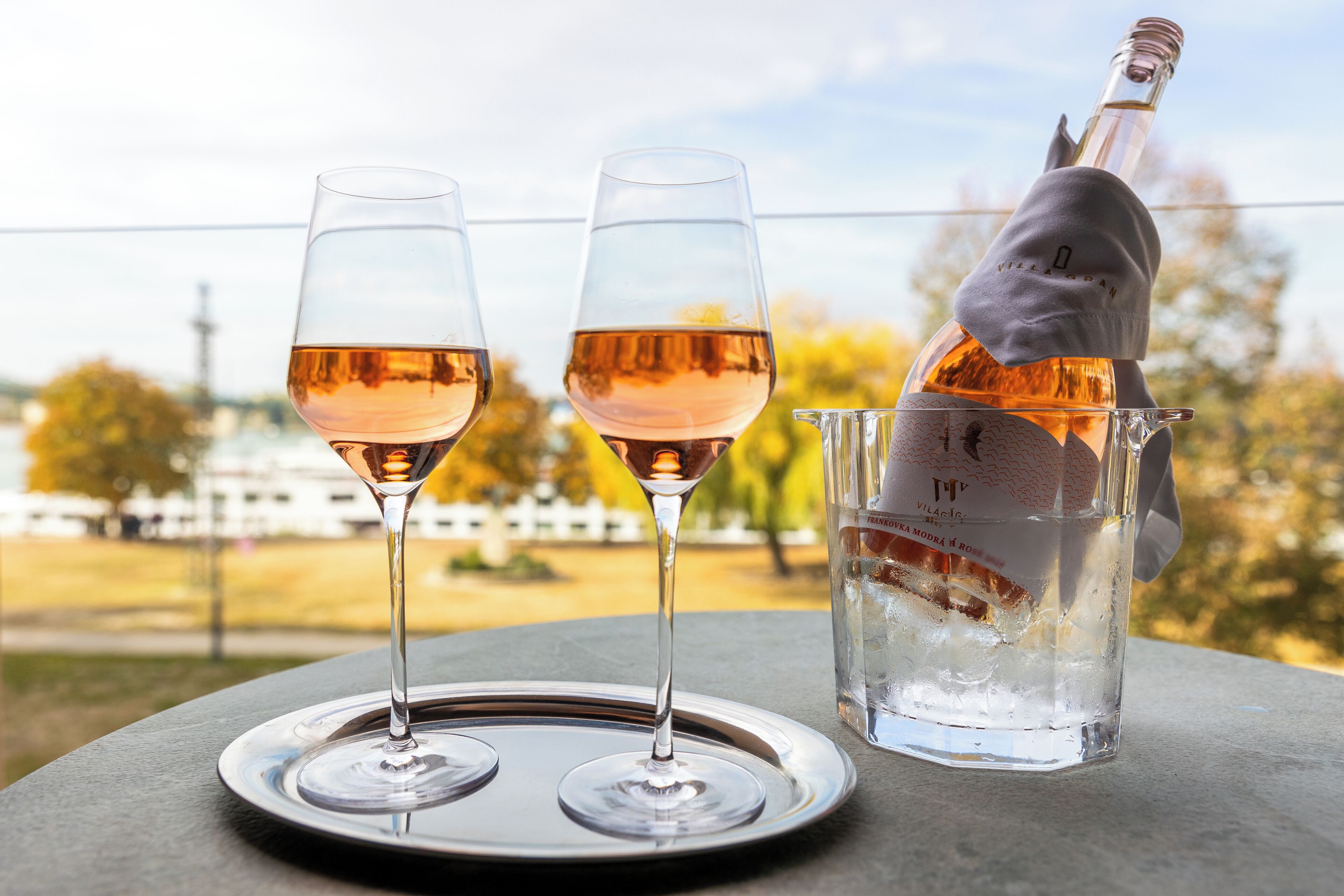Dos copas de vino rosado sobre una bandeja, una botella de vino rosado y una jarra con hielo, todo sobre una mesa al aire libre con vistas a árboles y un edificio al fondo.