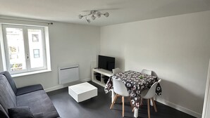 A modern living room with a sofa, a dining table with chairs, a TV, and a window. - Charming fully furnished T2 (Chavanay)
