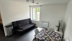 A compact room with a dining table, chairs, a sofa, a small trash bin, and a window. - Charming fully furnished T2 (Chavanay)