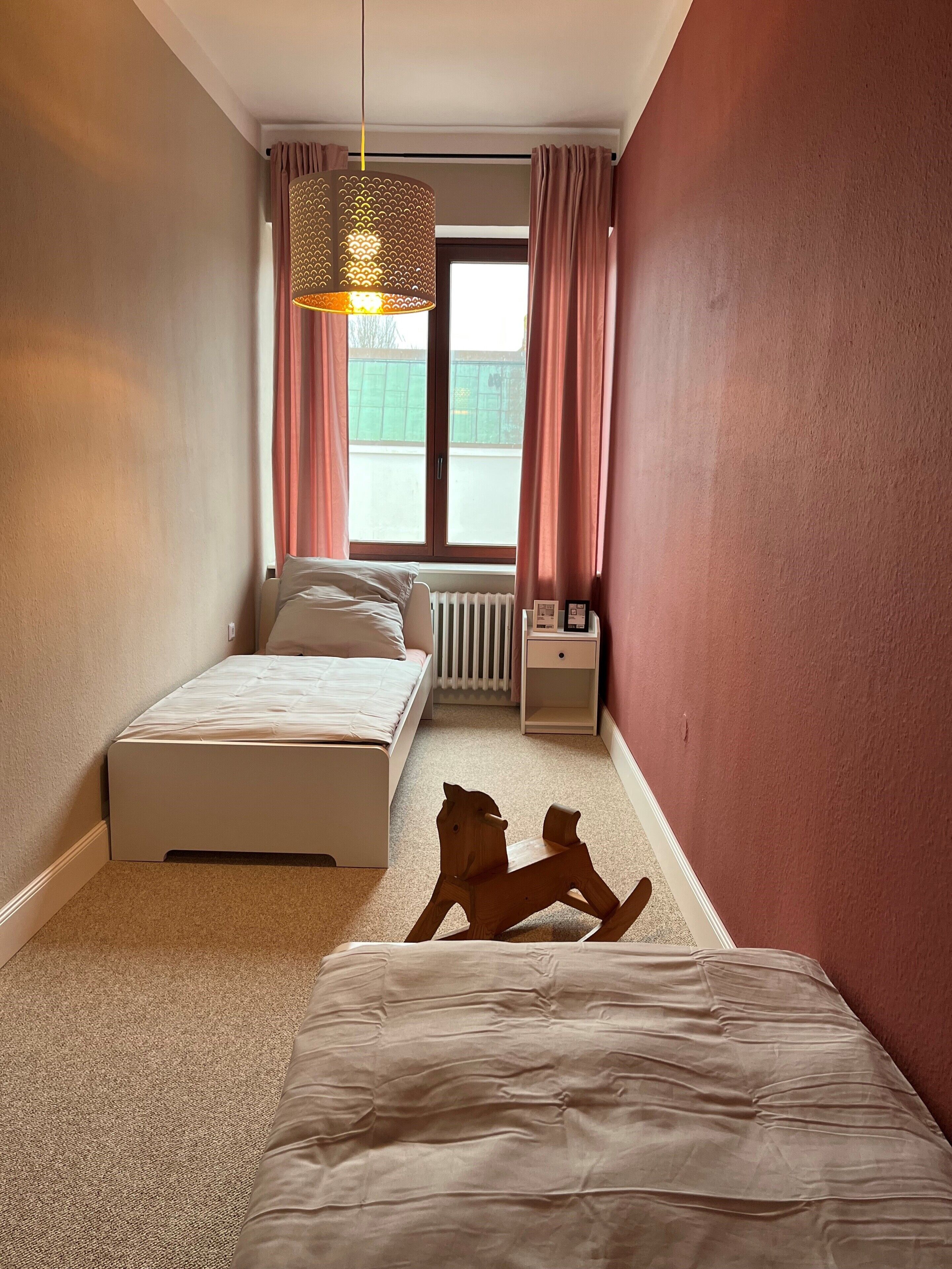 A bedroom with a bed, a radiator, a nightstand, a hanging light fixture, and a window with curtains.