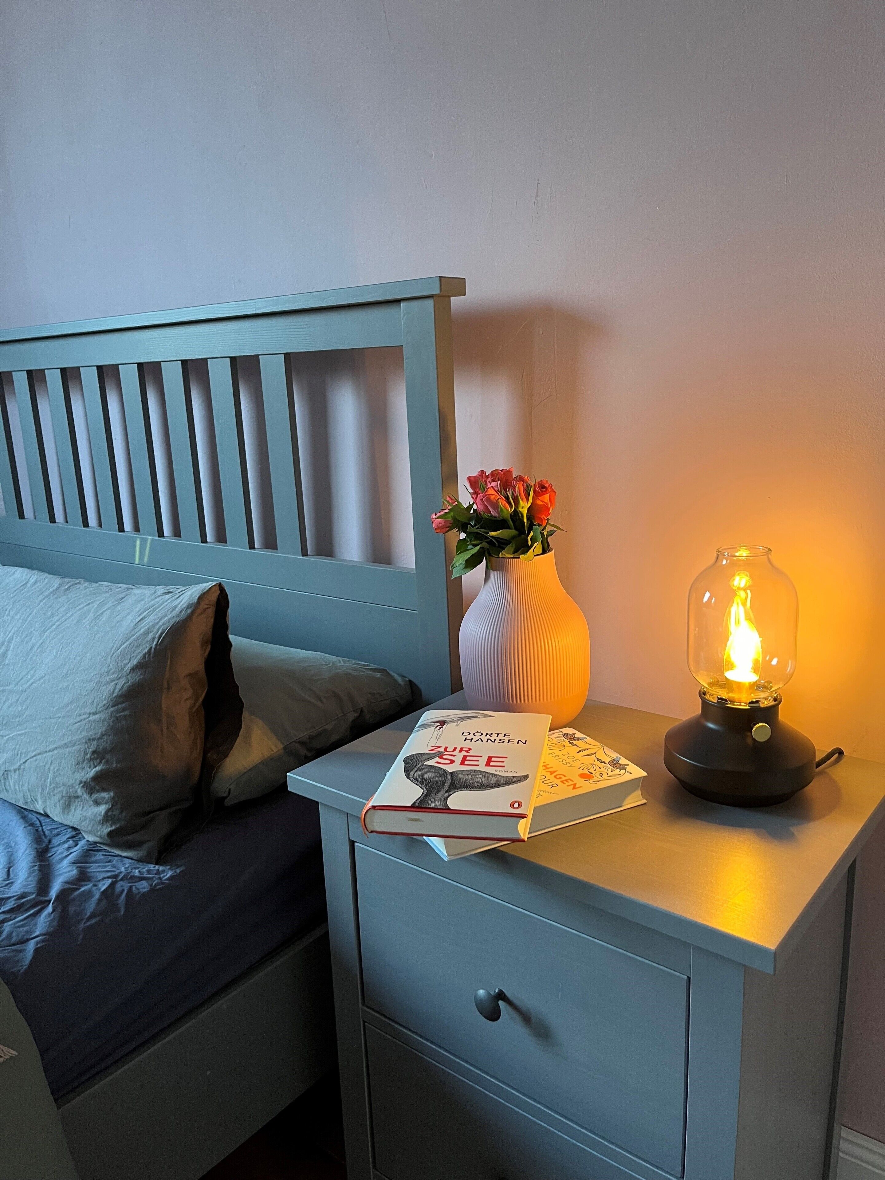 A bed with pillows, a bedside table with a book, a vase with flowers, and a lit lamp.