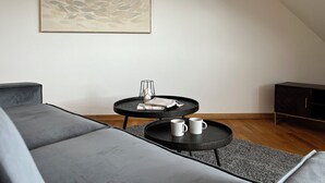 Een moderne woonkamer met een grijze bank, een klein rond tafeltje met twee witte mokken, een lijst met een abstract schilderij en een houten kast.