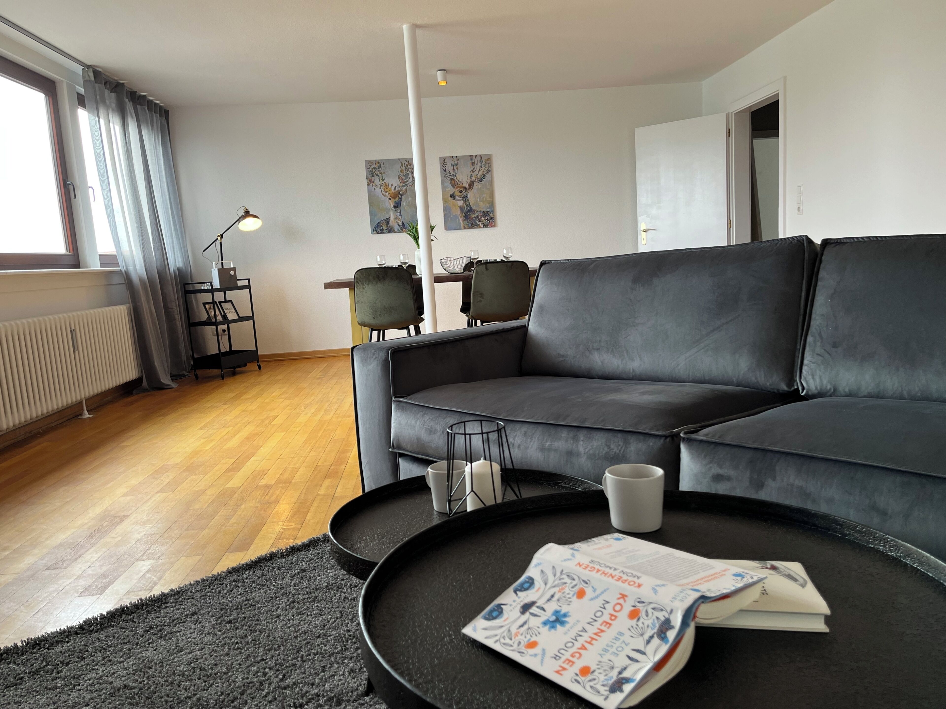 Ein modernes Wohnzimmer mit einem grauen Sofa, einem Couchtisch mit einer Zeitschrift und einem Essbereich im Hintergrund.