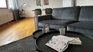 Een moderne woonkamer met een grijze bank, een bijzettafeltje met een tijdschrift, en een eethoek op de achtergrond.