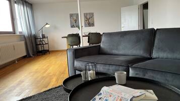 Ein modernes Wohnzimmer mit einem grauen Sofa, einem Couchtisch mit einer Zeitschrift und einem Essbereich im Hintergrund.
