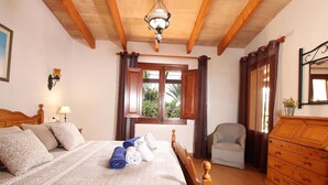 A bedroom with a bed, wooden headboard, a chair, a dresser, a window with curtains, and a ceiling fan. - Villa Can Tia in Pollensa with Pool, Garden & Wi-Fi (Illes Balears)