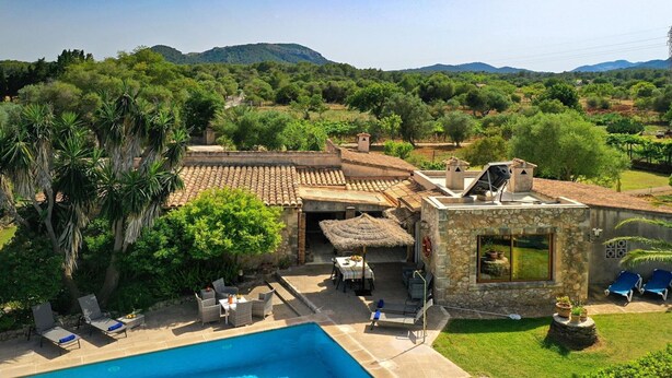 A poolside area with lounge chairs, a dining table, and a stone building with a view of the surrounding landscape. - Villa Can Tia in Pollensa with Pool, Garden & Wi-Fi (Illes Balears)