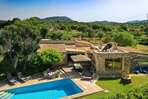 A poolside area with lounge chairs, a dining table, and a stone building with a view of the surrounding landscape. - Villa Can Tia in Pollensa with Pool, Garden & Wi-Fi (Illes Balears)