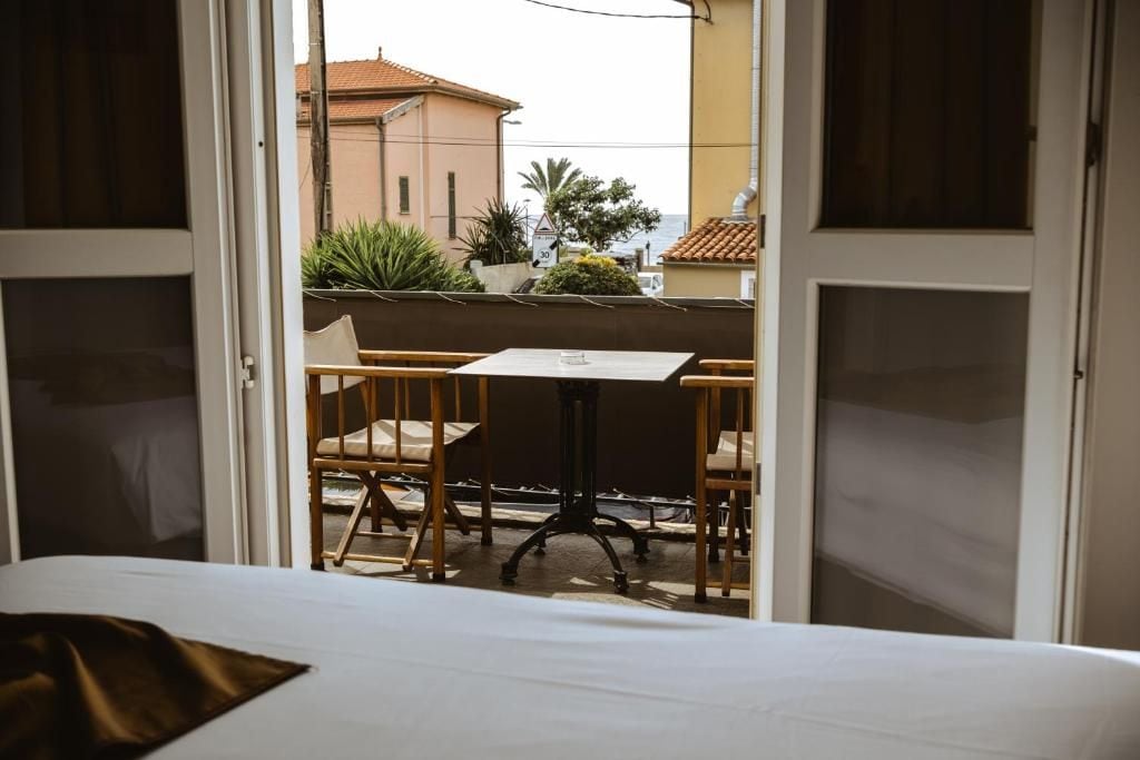 Une chambre d’hôtel avec un lit, une table et deux chaises. On peut voir depuis la fenêtre le balcon extérieur, avec des plantes et un b âtiment.