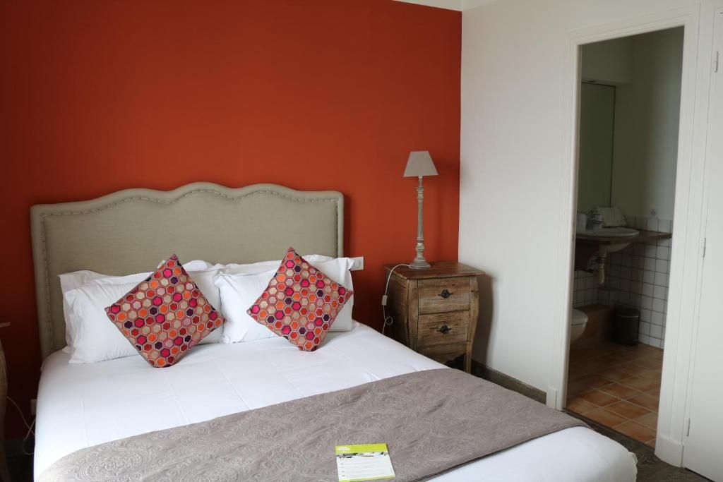 Quarto de hotel com cama, duas almofadas, mesa de cabeceira, luminária e banheiro visível por uma porta aberta.