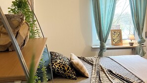 Quarto com cama de solteiro, janela com cortinas, mesa de cabeceira com abajur e prateleira na parede com plantas.