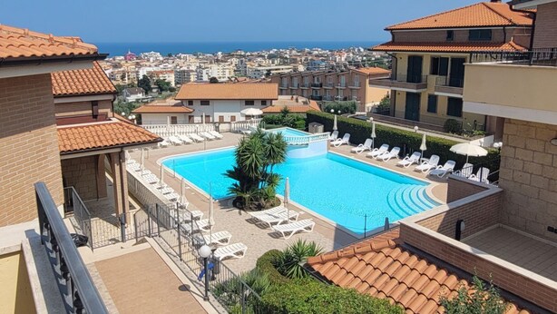 Uma piscina no terraço com espreguiçadeiras, vista para o mar e edifícios ao redor.