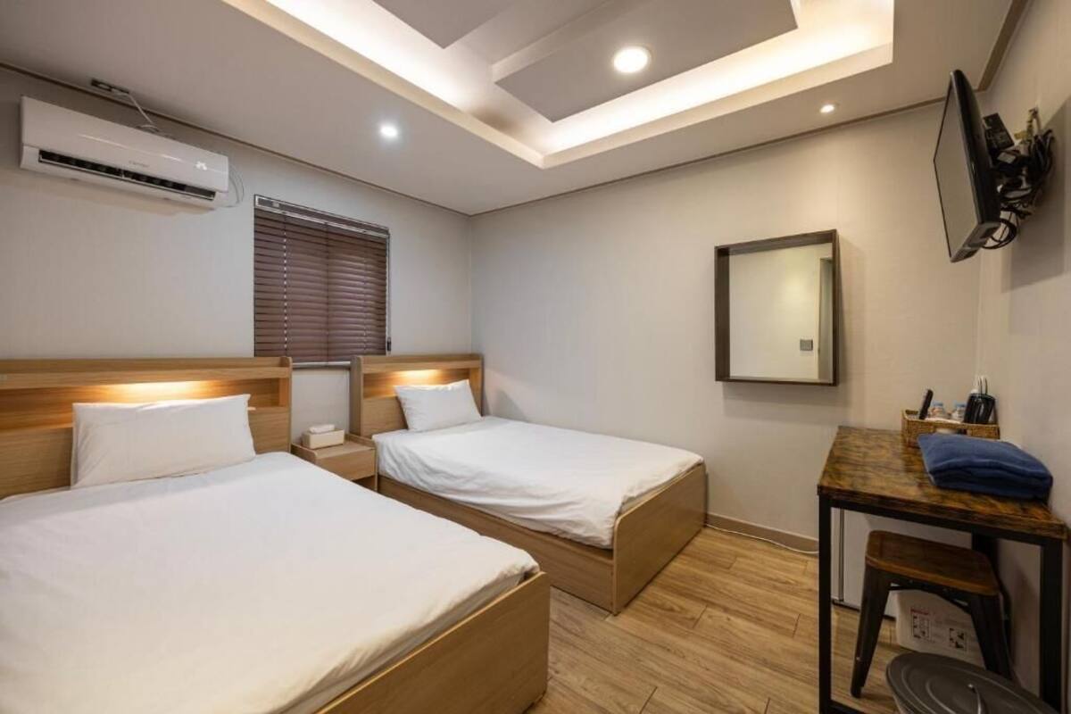 A hotel room with two beds, a desk, a chair, an air conditioning unit, a window with blinds, and a mirror on the wall.