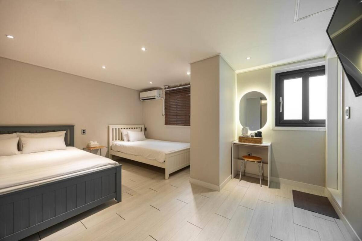 A bedroom with a large bed, a smaller bed, a desk with a chair, a mirror, and an air conditioning unit.