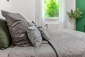 Ein Bett mit grauer Bettwäsche und gemusterten Kissen, ein Fenster mit Blick auf Grünflächen und eine Heizung.