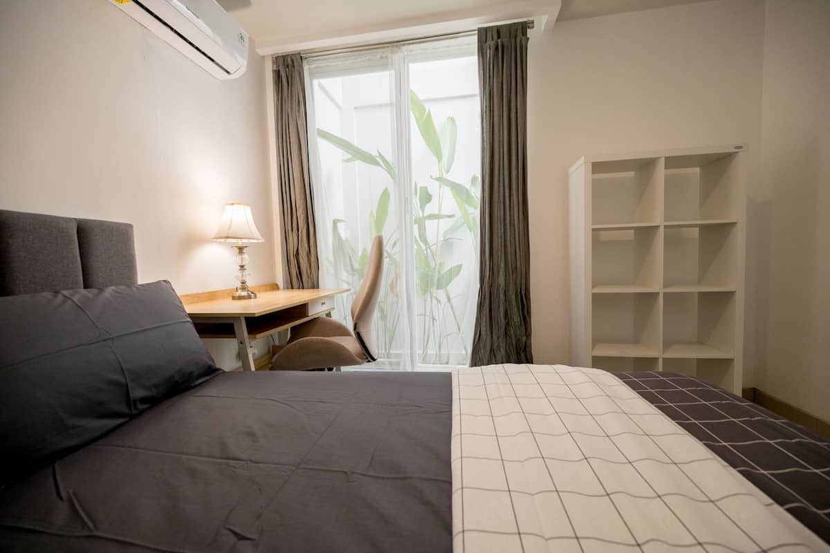 Hotellihuone, jossa on sänky, työpöytä lampun kanssa, ikkuna verhoilla ja seinään integroitu kirjahylly.