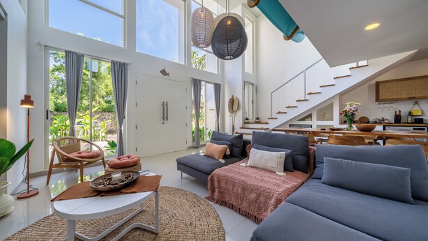 A spacious living area with a large sofa, a coffee table, and a dining table. There is a staircase leading to an upper level. - Park Homei (Pecatu)
