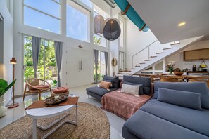 A spacious living area with a large sofa, a coffee table, and a dining table. There is a staircase leading to an upper level. - Park Homei (Pecatu)