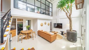 A modern living room with a sofa, dining table, and chairs, a large potted tree, and a staircase. - Park Homei (Pecatu)