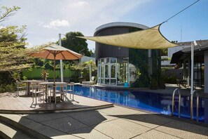 Ein Pool mit Sonnenschirm, Sitzgelegenheiten im Freien und ein Gebäude mit bogenförmiger Fassade.