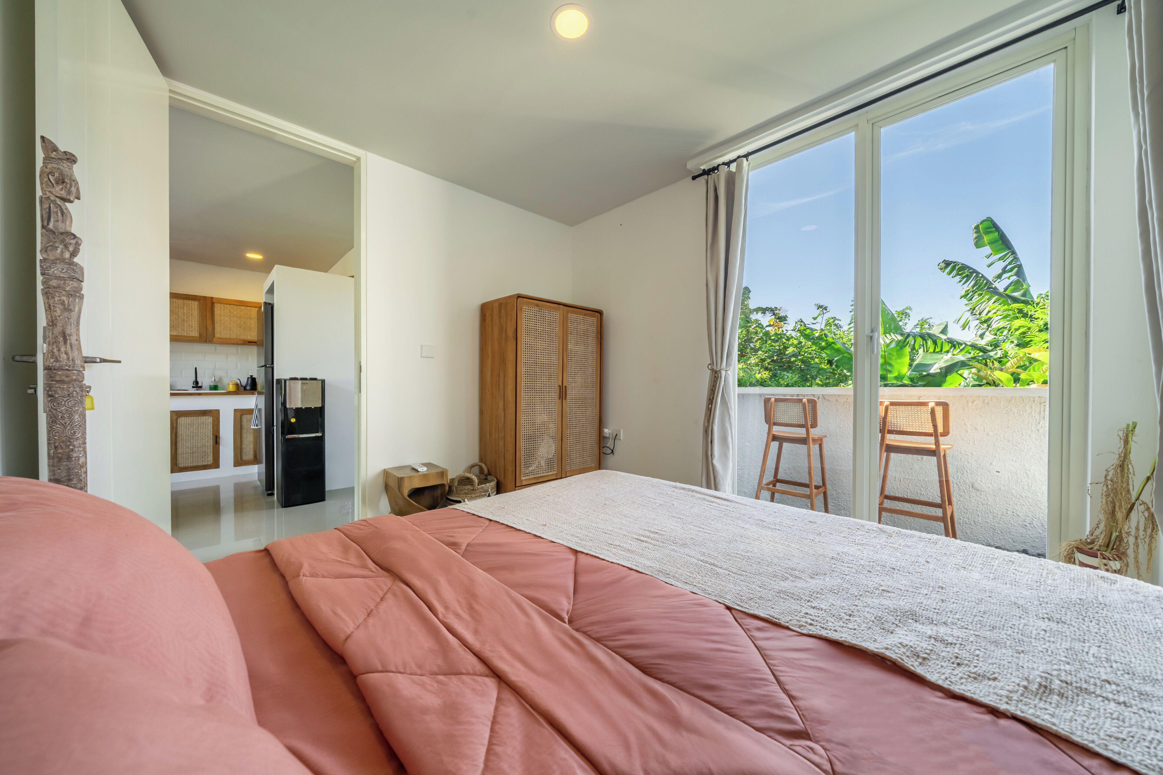 A bedroom with a bed, a wooden wardrobe, a kitchenette, and a balcony with chairs.