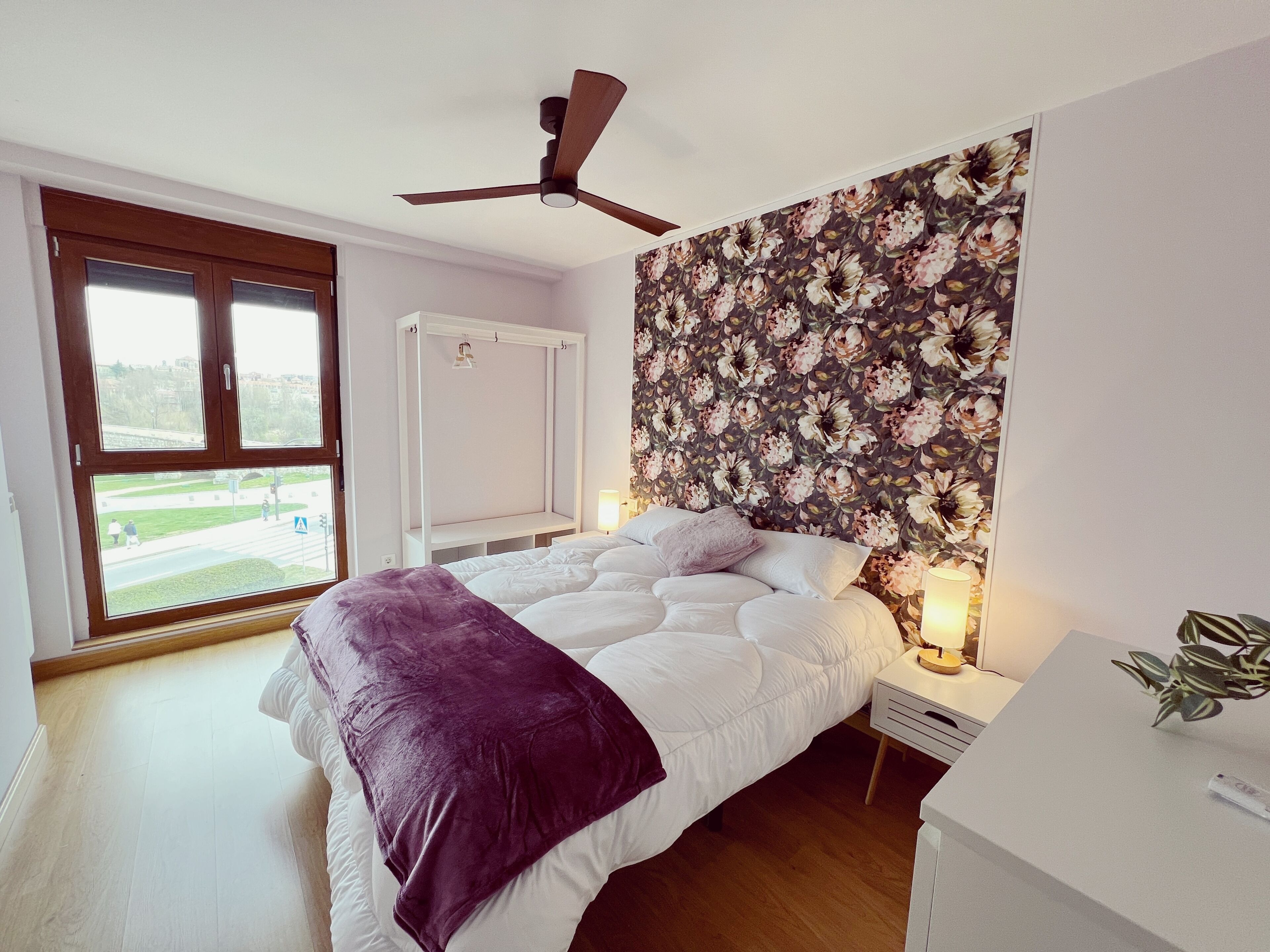 Un dormitorio con cama, ventilador de techo, ventana con vistas y papel tapiz floral.