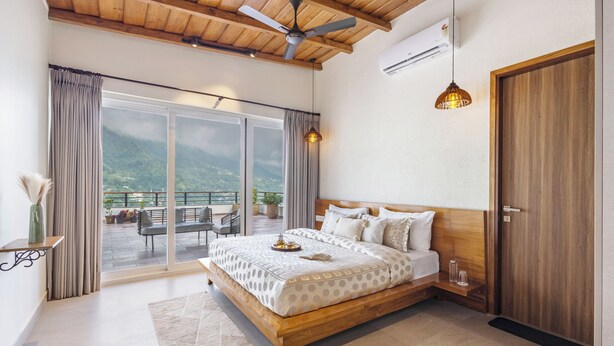 Un dormitorio con una cama grande, ventilador de techo, puerta de madera y vista a un balcón con mobiliario exterior.