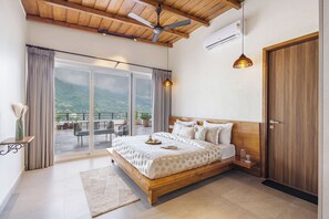A bedroom with a large bed, a ceiling fan, a wooden door, and a view of a balcony with outdoor furniture. - ELIVAAS Antarmann Casa Ember 2BHK   (Bhimtal)