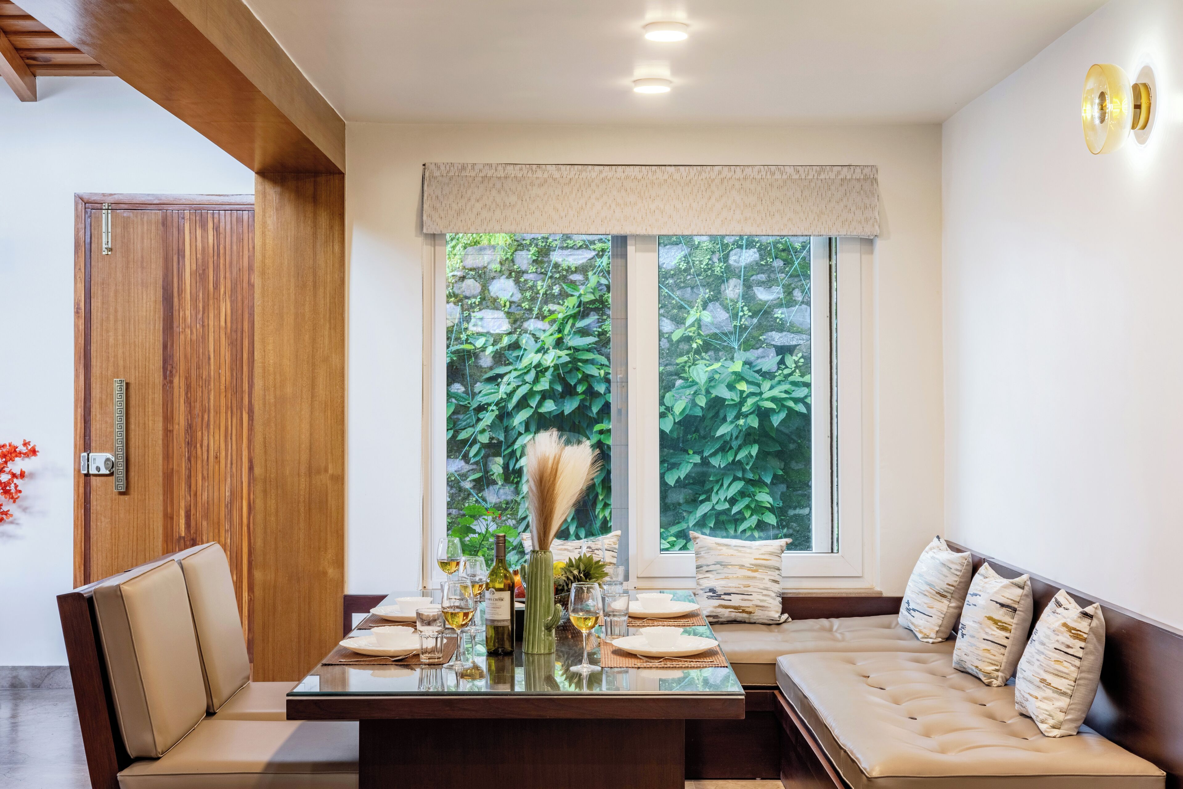Un comedor con mesa de madera dispuesta para cuatro, un banco acolchado y ventana con vista a follaje.