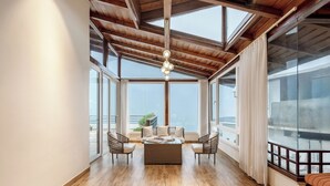 Una habitación amplia con techo alto, ventanas grandes, una zona de estar central con una mesa de centro de fibras naturales y un balcón con barandilla.