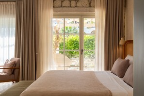 A bedroom with a large bed, curtains, a view of greenery, and a stone wall. - Fiora Alacati (Çeşme)