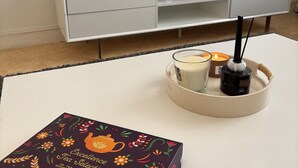 En moderne stue med et fladskærms-tv på væggen, et hvidt tv-bord med en fjernbetjening, en dekorativ bakke med stearinlys og en sort genstand, samt en æske tebreve på et hvidt bord.