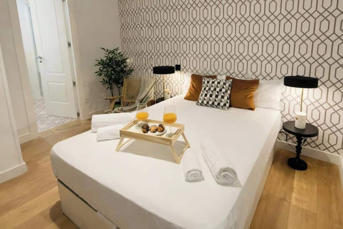 Ein ordentlich bezogenes Bett, ein Tablett mit Snacks, ein Nachttisch mit Lampe und eine Wand mit geometrischem Muster.