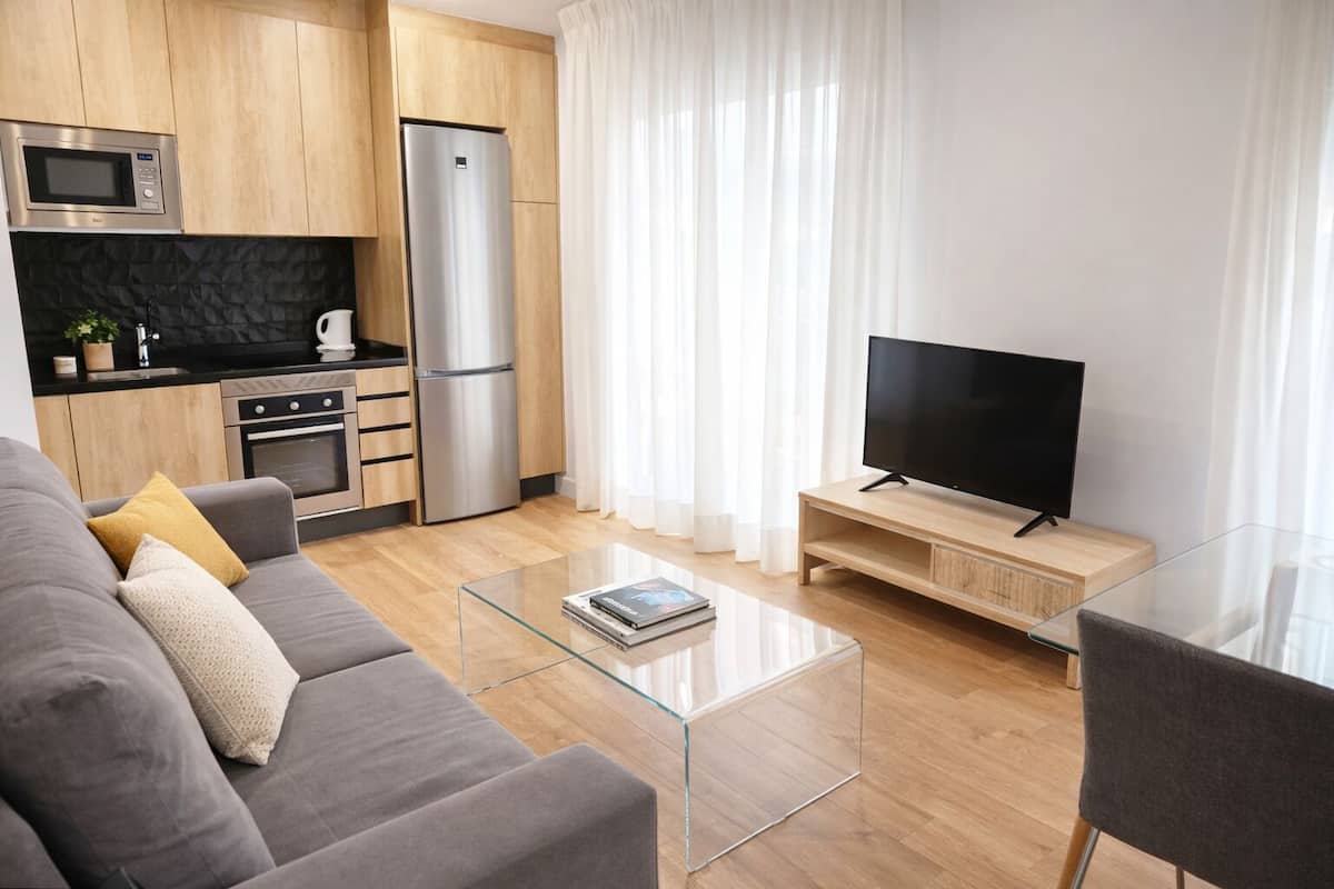 Ein modernes Wohnzimmer mit einer Couch, einem Glastisch, einem Flachbildfernseher, einer Küchenzeile mit Kühlschrank, Backofen und Mikrowelle sowie einem Fenster mit durchsichtigen Vorhängen.