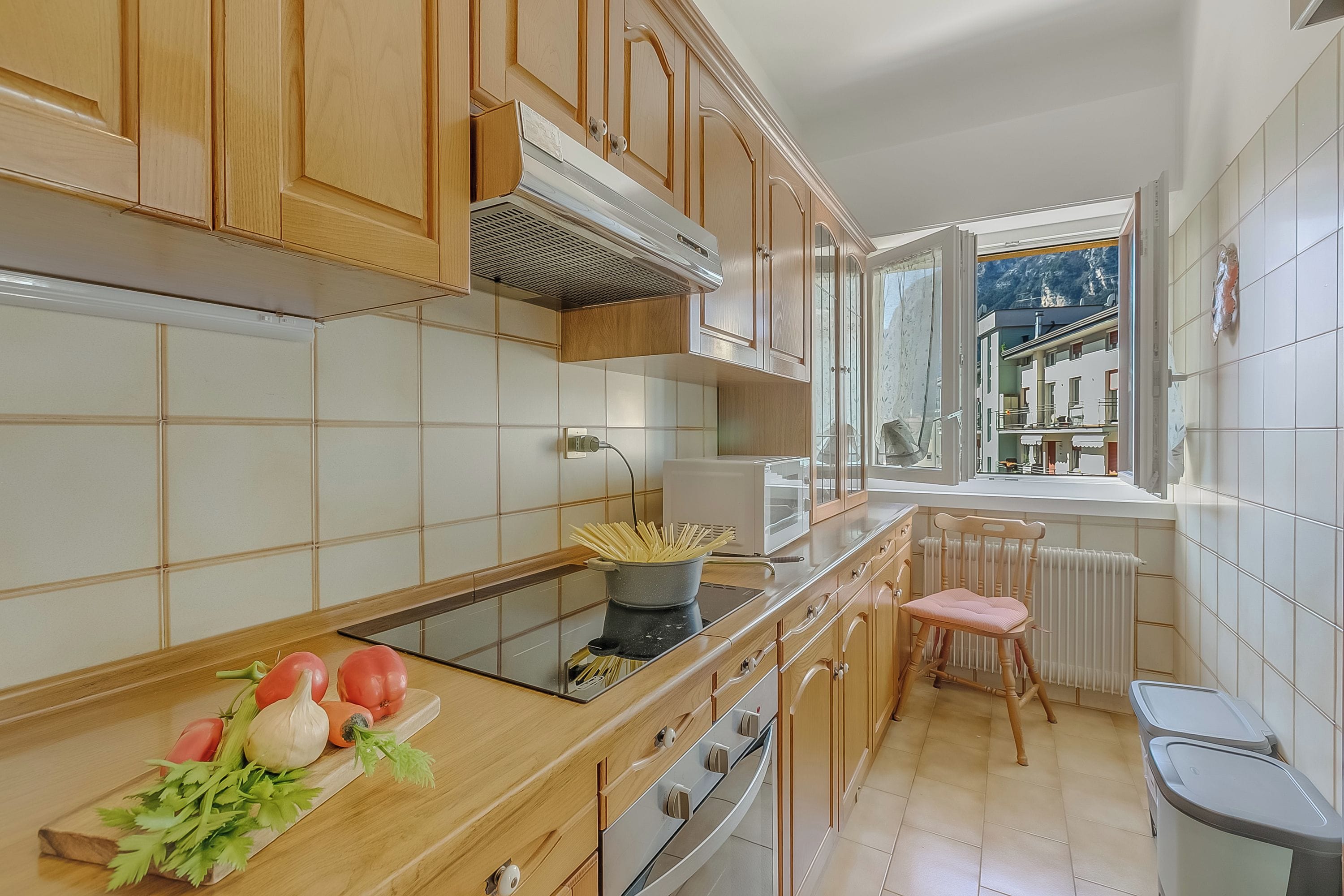 Una cucina con mobili in legno, una finestra che si affaccia su edifici e un bancone con verdure.