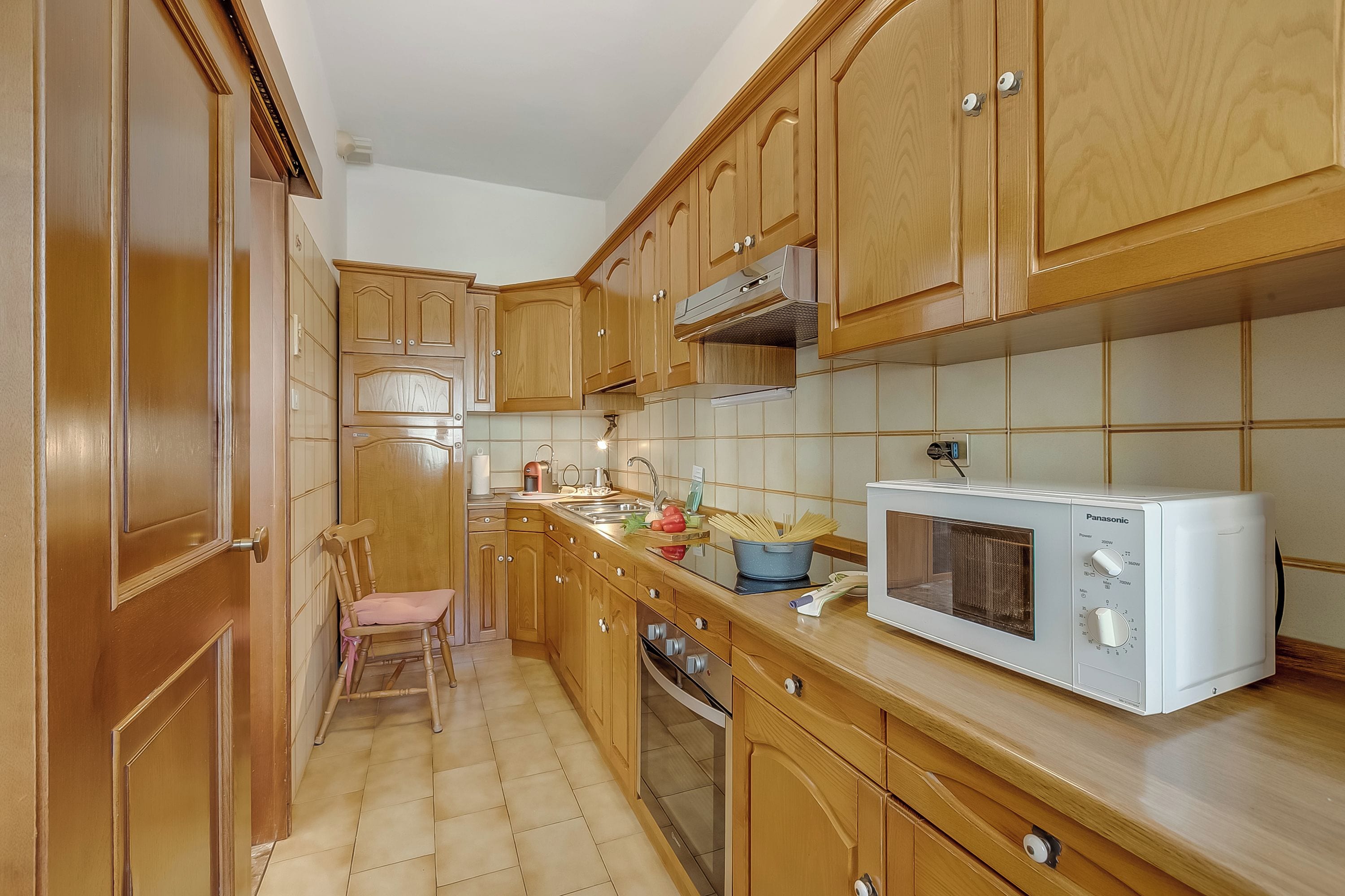 A kitchen with wooden cabinets, a microwave, a sink, and a chair.
