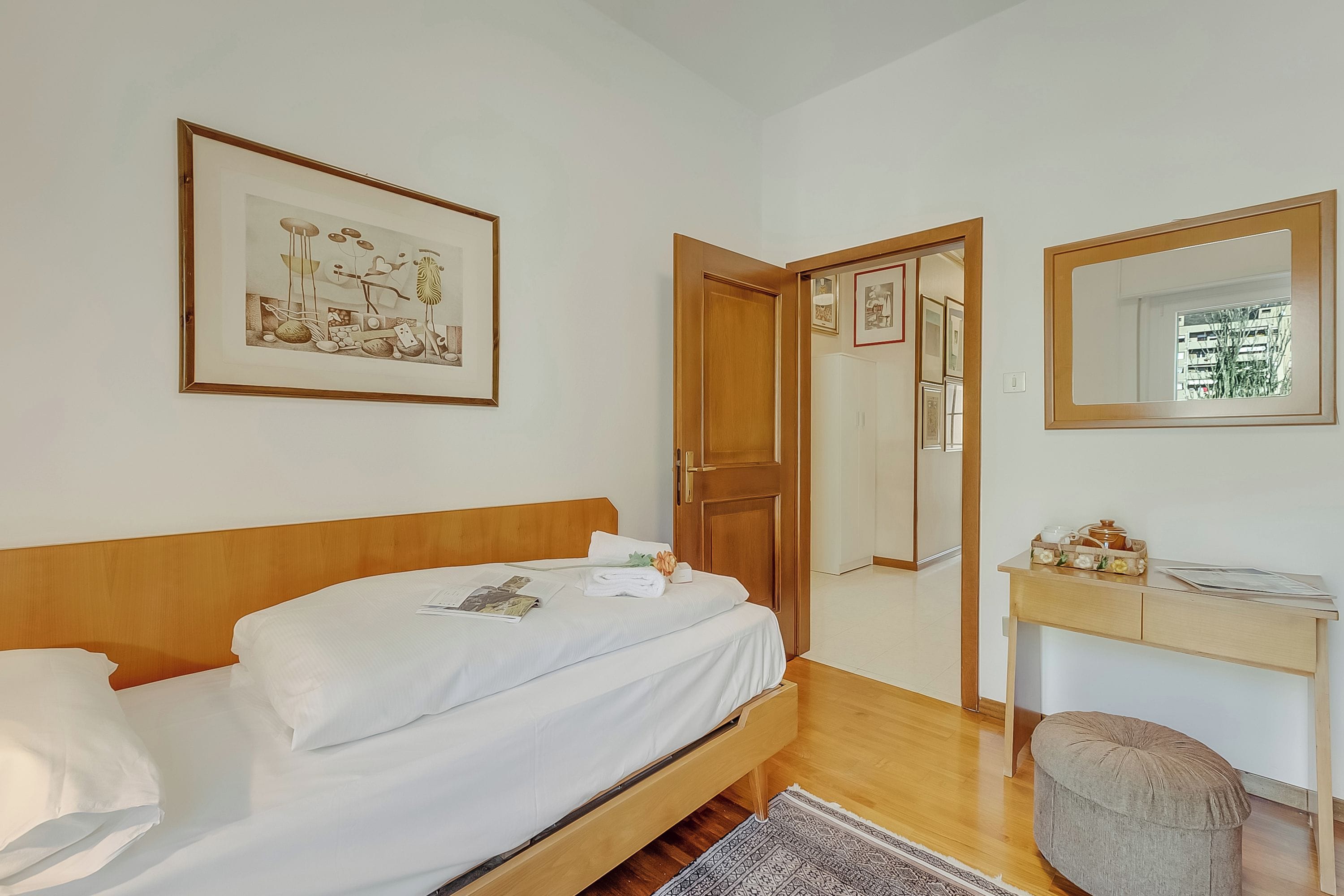 Un lit bien fait, avec du linge de lit blanc, une tête de lit en bois, un tableau encadré au mur, un miroir au-dessus d’un bureau et une porte donnant sur une autre pièce.