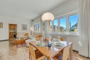 A modern living room with a dining area, large windows, and a view of mountains. - Rosa Apartment, Balcony and AC (Riva del Garda)
