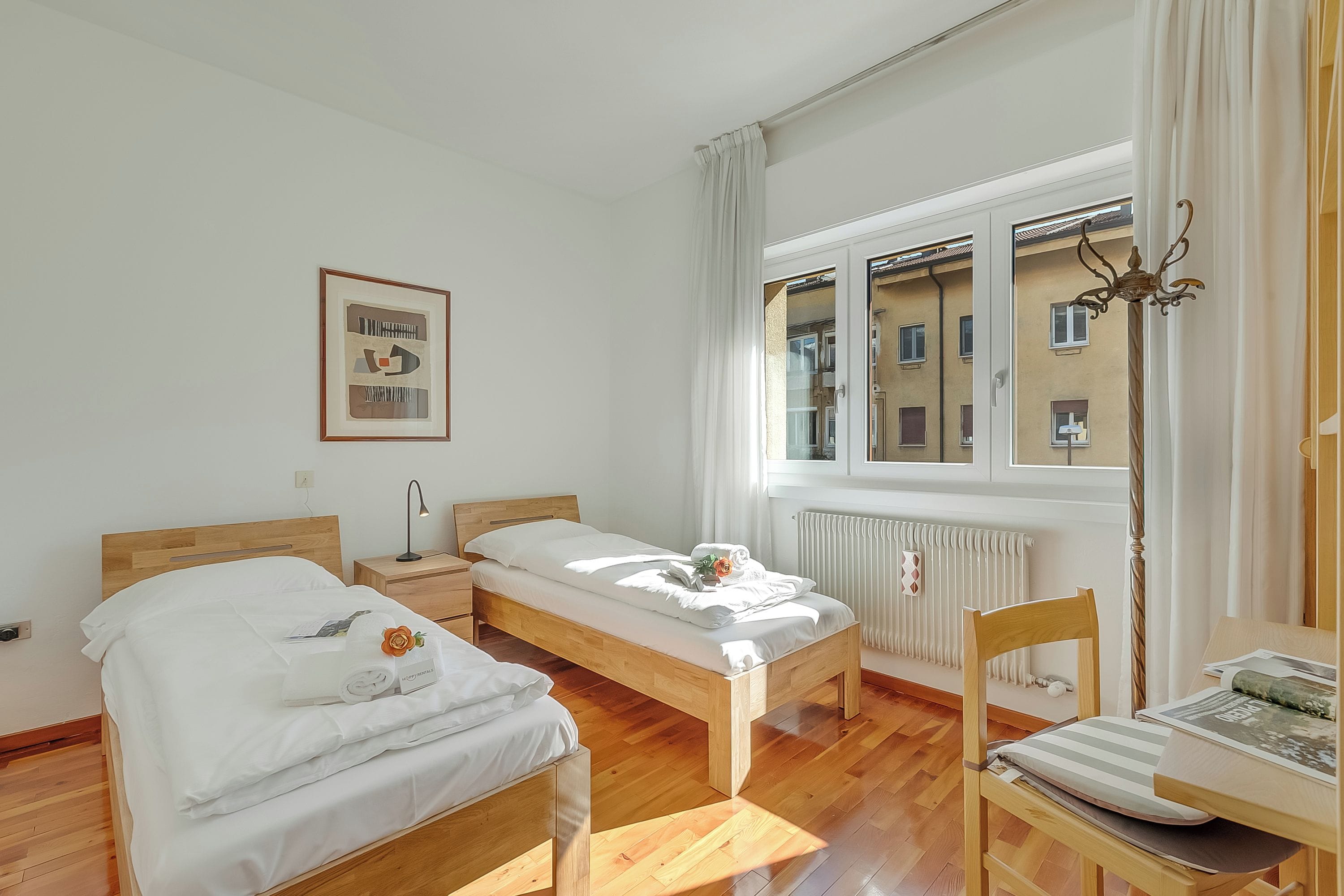A room with two wooden beds, a small table, a chair, a radiator, and a window with curtains.