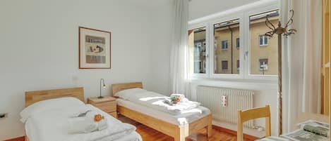 A room with two wooden beds, a small table, a chair, a radiator, and a window with curtains.