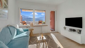 A living room with a sofa, a TV, a small table with a bottle and glasses, and a window with a view of mountains. - Bella Vista Apartment (Riva del Garda)