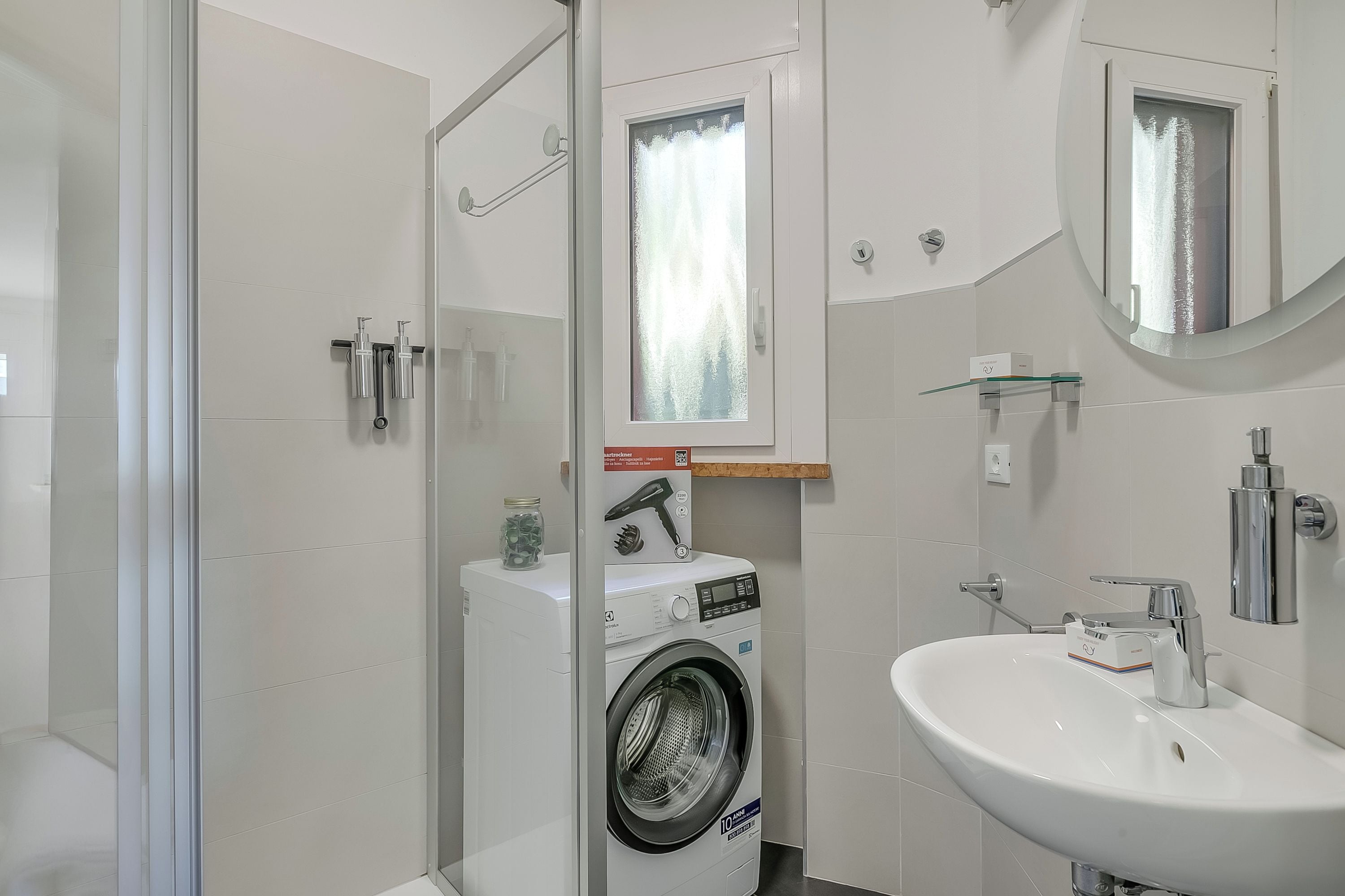 A modern bathroom with a glass shower enclosure, a built-in washing machine, and a sink with a mirror above.