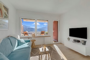 A living room with a sofa, a TV, a small table with a bottle and glasses, and a window with a view of mountains. - Bella Vista Apartment (Riva del Garda)